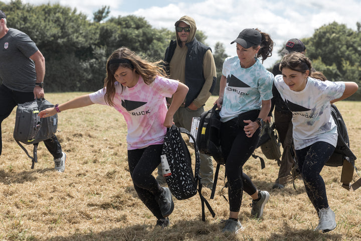 Three women in athletic gear run across a grassy field, carrying backpacks, during an outdoor event.