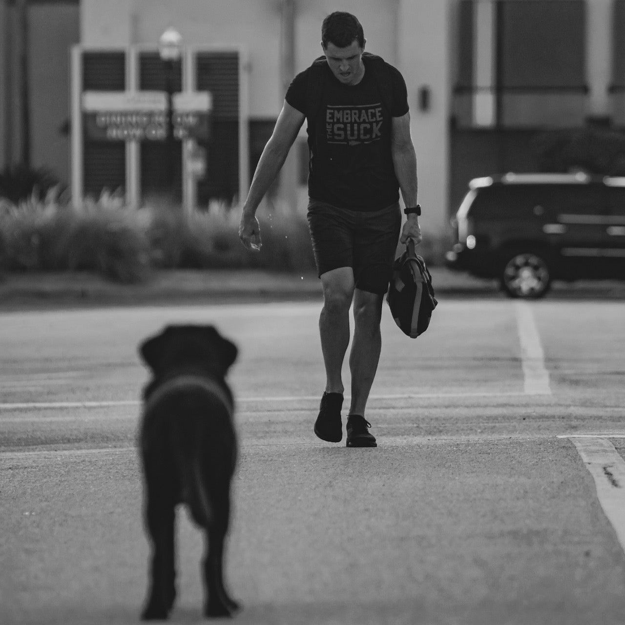 A man walks toward a dog on a street, carrying a bag, in black and white—wearing the Embrace the Suck Tee - Tri-BlendX, perfect for reminding us to stay resilient during tough times.
