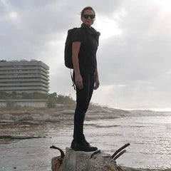 Person wearing all-black rugged gear and backpack standing on rocky outcrop by rough ocean under cloudy sky