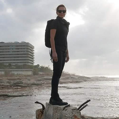A person wearing sunglasses stands on a rock by the coast, dressed in black with a GORUCK GR1 USA - Dyneema rucksack. The sky is overcast, and a modern building is visible in the background.