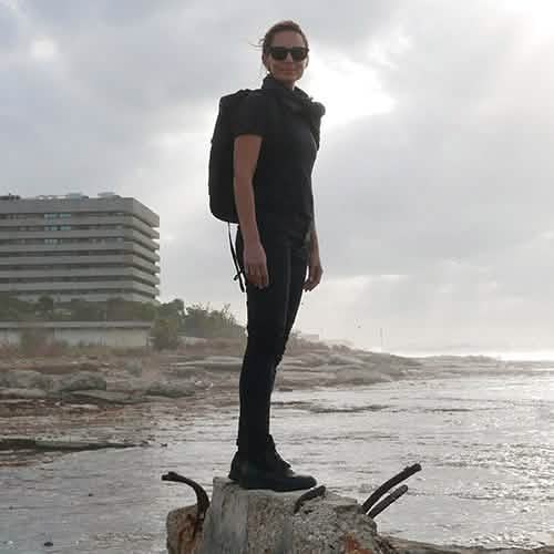 A person wearing sunglasses stands on a rock by the coast, dressed in black with a GORUCK GR1 USA - Dyneema rucksack. The sky is overcast, and a modern building is visible in the background.