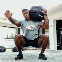 A man in a gray T-shirt featuring an American flag design is squatting outdoors, demonstrating core stability. He holds a black, round Sand Medicine Ball from GORUCK on his right shoulder, with his left arm extended forward. Dressed in black shorts and sneakers, he illustrates how this workout is suitable for a home gym setup.