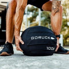 A person dressed in athletic apparel crouches outdoors to lift a black Sand Medicine Ball labeled GORUCK 100#, highlighting the veins and tattoos on their arm. This home gym-friendly ball is perfect for boosting core stability during workouts.
