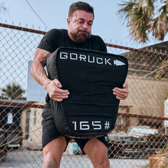 Man lifting a black 165-pound GORUCK weighted bag outdoors near chain-link fence