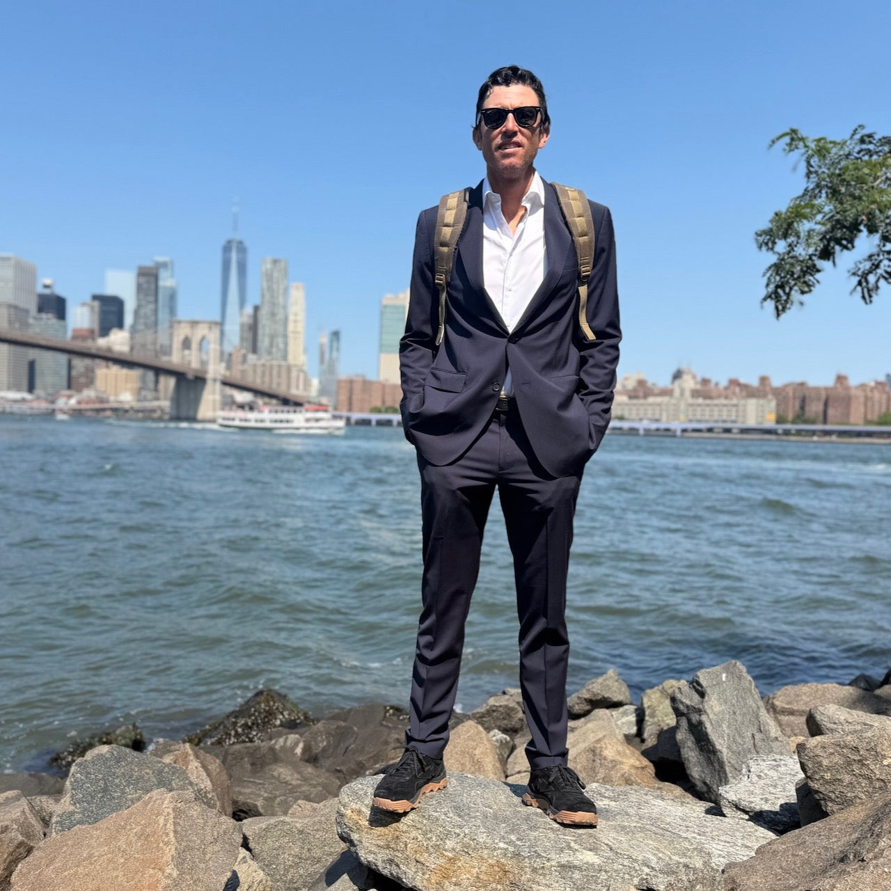 A man in a suit with the GR1 USA - Heritage Waxed Canvas rucksack stands on rocks by the water, city skyline and bridge in the background.