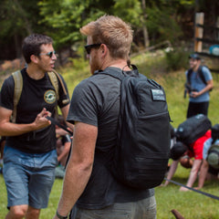 Two men with Bullet Ruck USA Throwback backpacks featuring MOLLE webbing and sunglasses talk outdoors, while others exercise on the grass in the background.
