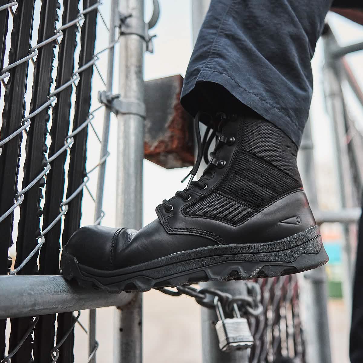 A close-up showcases a person wearing the MACV-2 Safety Boot - High Top from GORUCK as they scale a metal fence. The boot is firmly planted on a metal bar, with a chain and lock visible nearby, perfectly complementing dark pants designed to Special Forces standards for the ultimate stealthy ensemble.