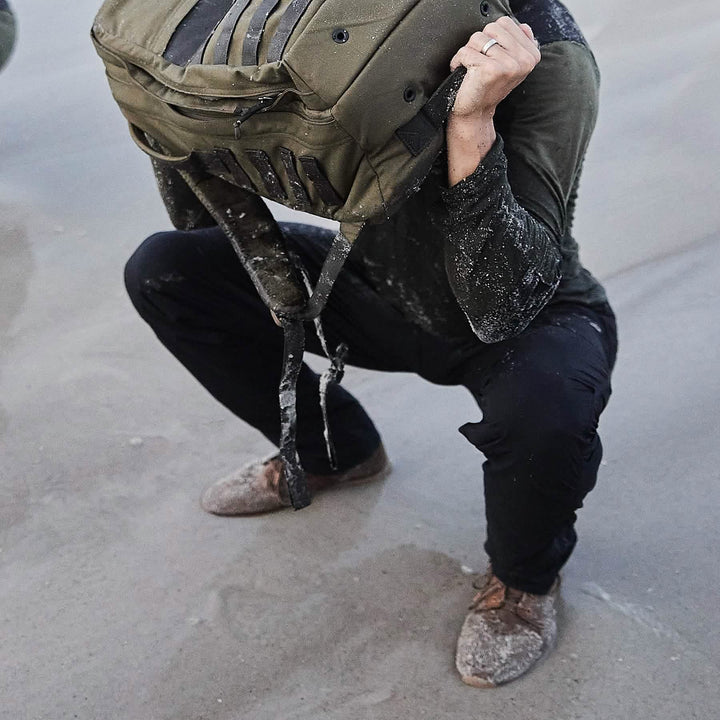 A person squats on a sandy surface, lifting a large bag overhead. Dressed in dark, sand-covered MACV-1 Hi-Speed - Mid Top boots by GORUCK and an outfit that highlights their determined posture and engaged muscles, the scene showcases strength paired with lightweight agility.