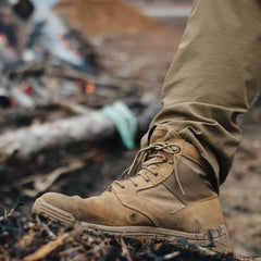 A person wearing the GORUCK MACV-2 Mid Top boots in Coyote, featuring a triple compound outsole, is standing on charred ground with olive green pants, against a blurred background of logs and flames.