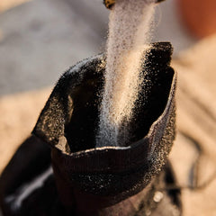 A close-up shot of fine sand cascading into a black fabric bag, ideal for crafting GORUCK Sand Medicine Balls. The sunlight accentuates the grains as they descend, creating a textured and dynamic visual against the open top of the bag, perfect for enhancing core stability in your home gym workouts.
