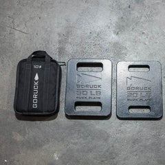 Three GORUCK items are positioned on the rough concrete floor: a sleek black rectangular 10-pound sandbag equipped with a handle, and two robust Sand Ruck Plates from GORUCK, labeled 30 LB and 20 LB, each featuring convenient cut-out handles. Discover the dependable performance of GORUCK gear backed by a Lifetime Guarantee.