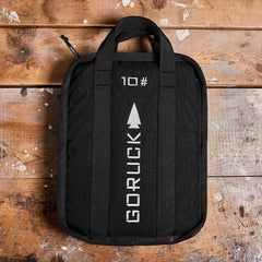 A Sand Ruck Plates bag by GORUCK, featuring two handles, rests on a worn wooden surface. The bag displays a white arrow logo with the text 10 GORUCK printed vertically, and it is backed by a Lifetime Guarantee for enduring adventures.