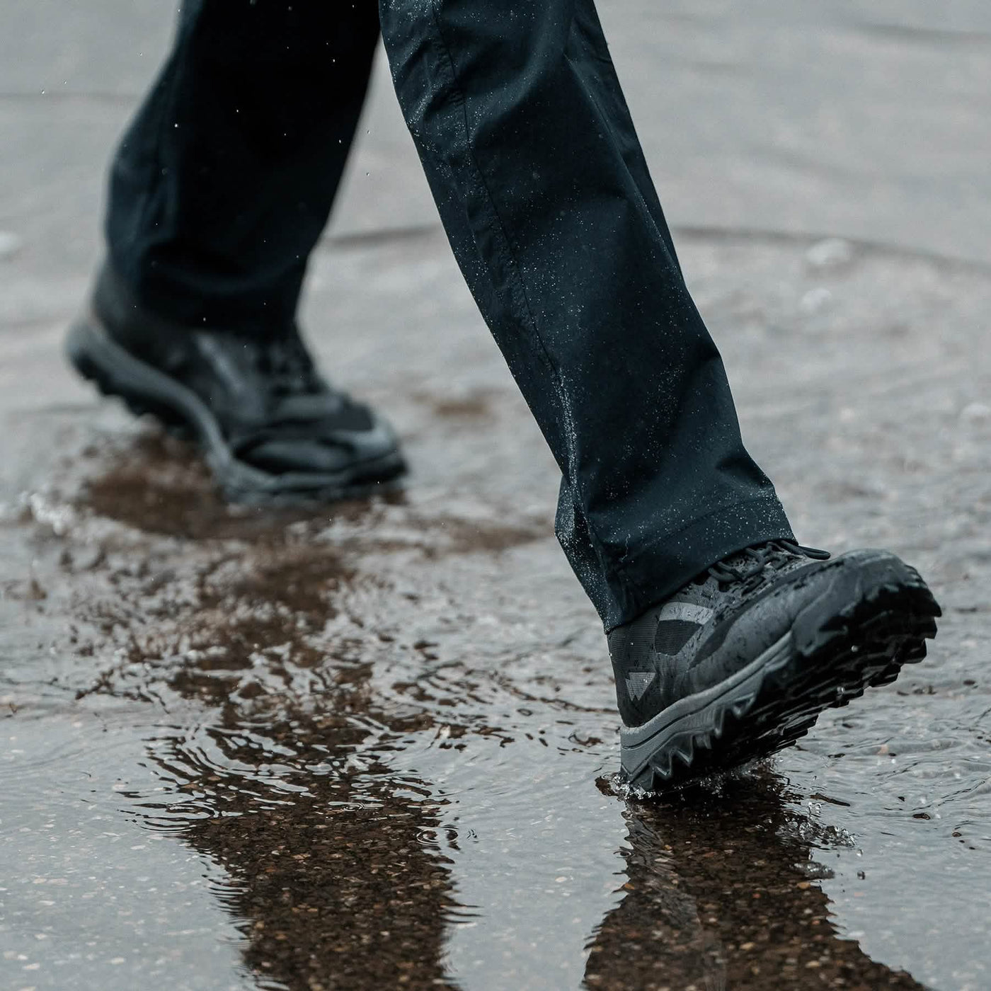 Clad in GORUCK’s Mackall boots in Black + Forged Iron, a person strides across a wet surface with ease, sending splashes around their dark pants as if they are executing a Special Forces training drill. The slightly blurred background highlights the dynamic motion through the puddle.