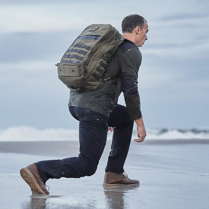 On a wet, sandy beach, a person with a large backpack kneels wearing a long-sleeve shirt, dark pants, and GORUCK's MACV-1 Hi-Speed - Mid Top rucking boots. Waves roll in the background beneath a cloudy sky.