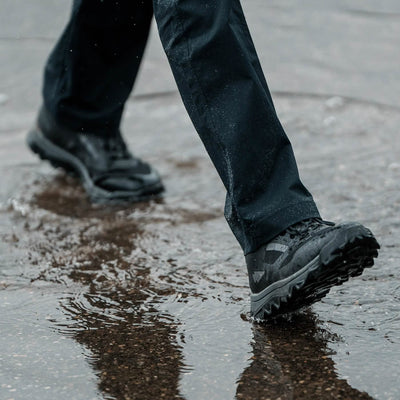 Clad in GORUCK’s Mackall boots in Black + Forged Iron, a person strides across a wet surface with ease, sending splashes around their dark pants as if they are executing a Special Forces training drill. The slightly blurred background highlights the dynamic motion through the puddle.