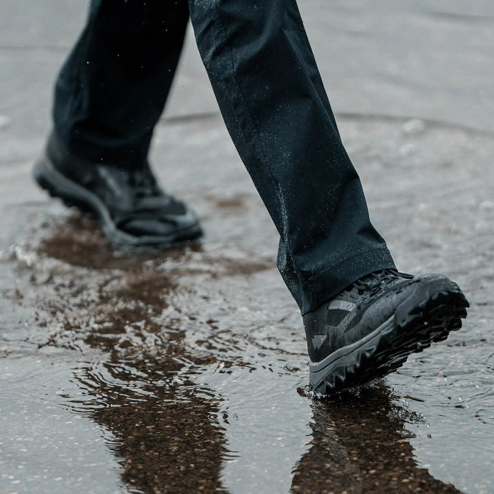 Clad in GORUCK’s Mackall boots in Black + Forged Iron, a person strides across a wet surface with ease, sending splashes around their dark pants as if they are executing a Special Forces training drill. The slightly blurred background highlights the dynamic motion through the puddle.