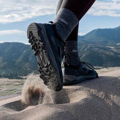 Close-up of a person wearing GORUCK's Mackall - Black + Forged Iron shoes paired with gray socks, stepping in sand on a dune. Sand is being kicked up by the shoes, with scenic mountains and a cloudy sky in the background.