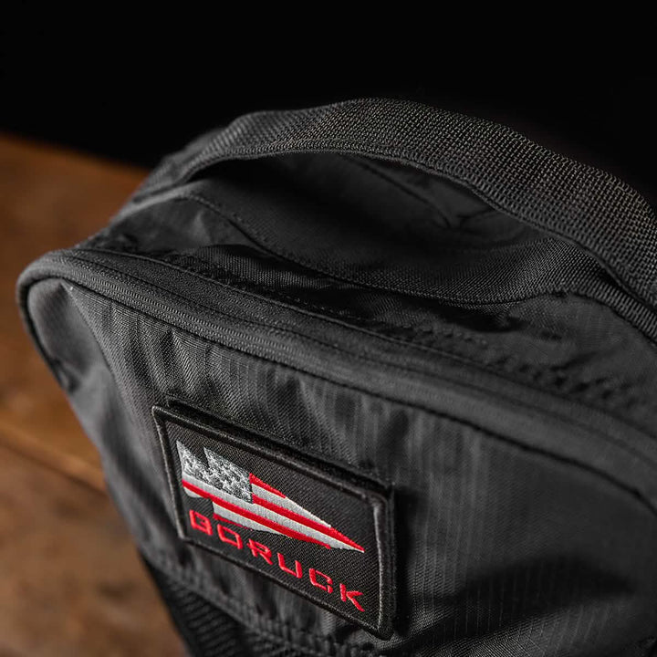 Close-up of a Shoe Bag - Ripstop ROBIC® in black, adorned with a patch that features a stylized American flag and the text GORUCK in red. The fabric texture is visible, accentuating its water-resistant quality, and the bag is set against a dark background with part of a wooden surface visible.