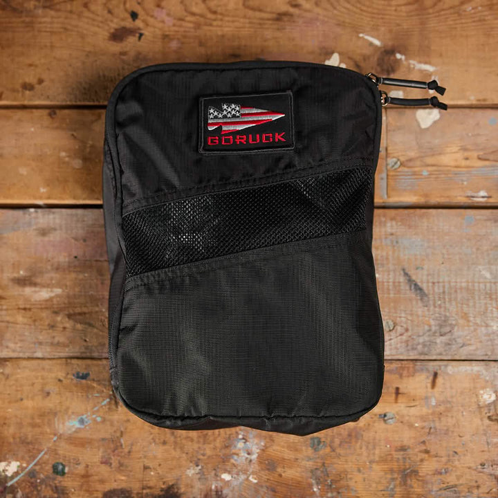 A black GORUCK Shoe Bag - Ripstop ROBIC® rests on a weathered wooden surface, featuring a front mesh pocket and dual zippers for added practicality. A patch displaying an American flag signifies its durability as a water-resistant bag. Paint splatters on the wood enhance the scene's rugged charm.