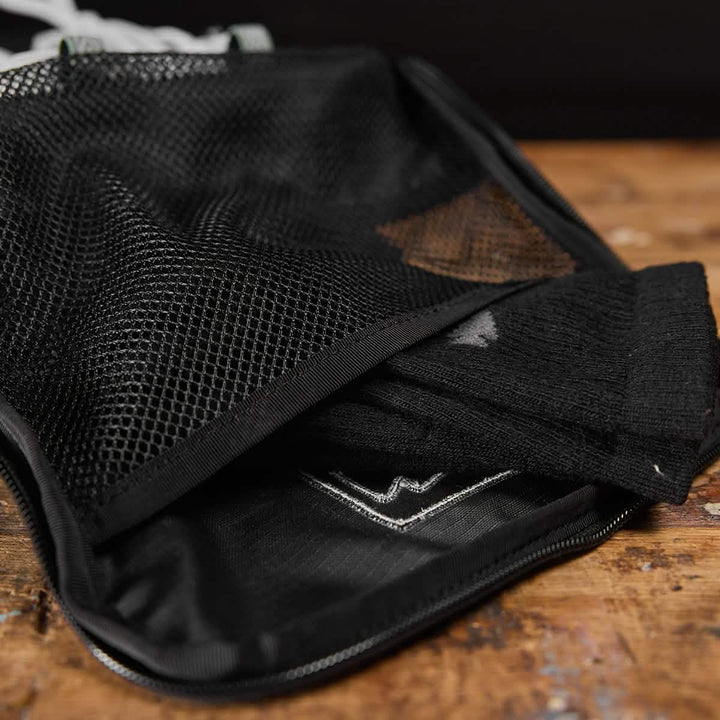 A pair of black sports socks is visible from an open black mesh pouch resting on a wooden surface. This GORUCK Shoe Bag, made with water-resistant Ripstop ROBIC® material, ensures durability and features a subtle logo design on the socks. Ideal for travel, this functional shoe bag comes with a lifetime guarantee.