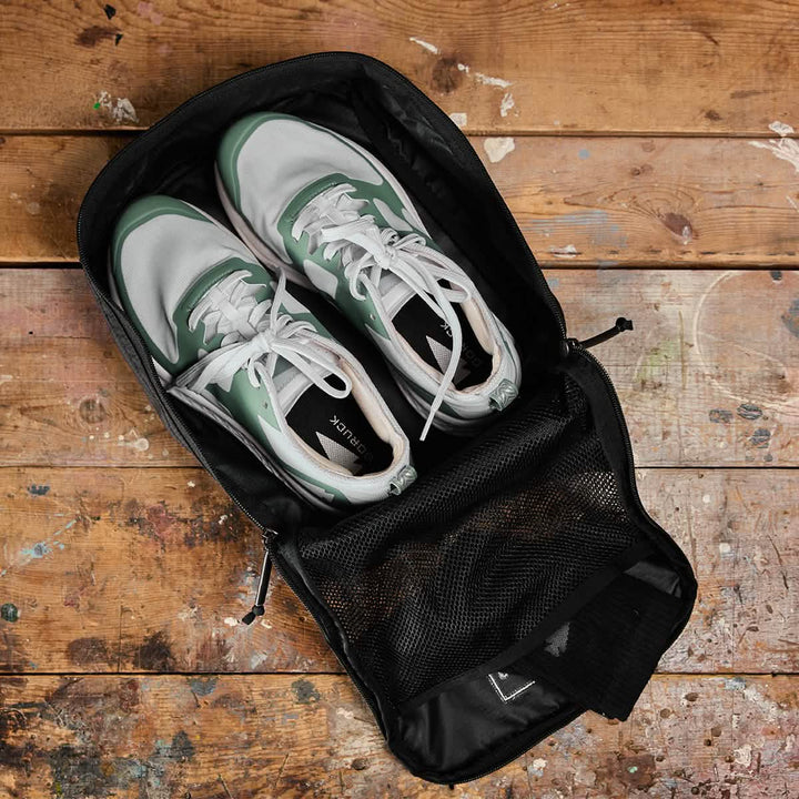 A pair of water-resistant white and green sneakers are tucked into an open, black, functional GORUCK Shoe Bag made of Ripstop ROBIC® fabric, featuring a mesh pocket on the inside flap. They are all resting on a rustic wooden floor with paint splatters.