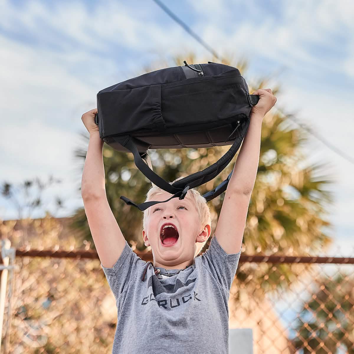A child with short blonde hair joyfully holds the GORUCK KR1, a black kids rucksack with padded straps, above their head. They are wearing a gray shirt and standing in front of a chain-link fence, with a tree and blue sky in the background.