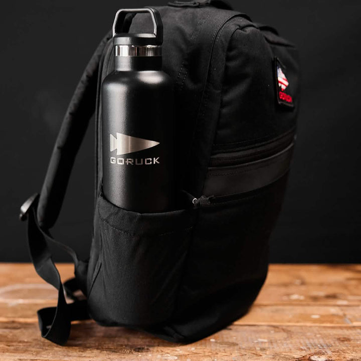 A black GORUCK KR1 kids rucksack rests on a wooden surface against a dark background. A black water bottle with the GORUCK branding is tucked in the side pocket. The backpack, ideal for school supplies, includes padded straps and features a small patch with a flag design.