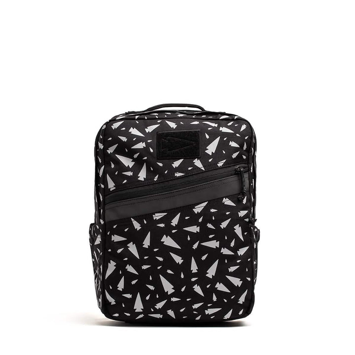 The KR1 - Spearhead Splatter by GORUCK is a kids' rucksack in black, adorned with a white paper airplane pattern. It features a front diagonal zipper pocket, a top handle for easy carrying, and textured fabric design that includes side pockets, perfect for school supplies.