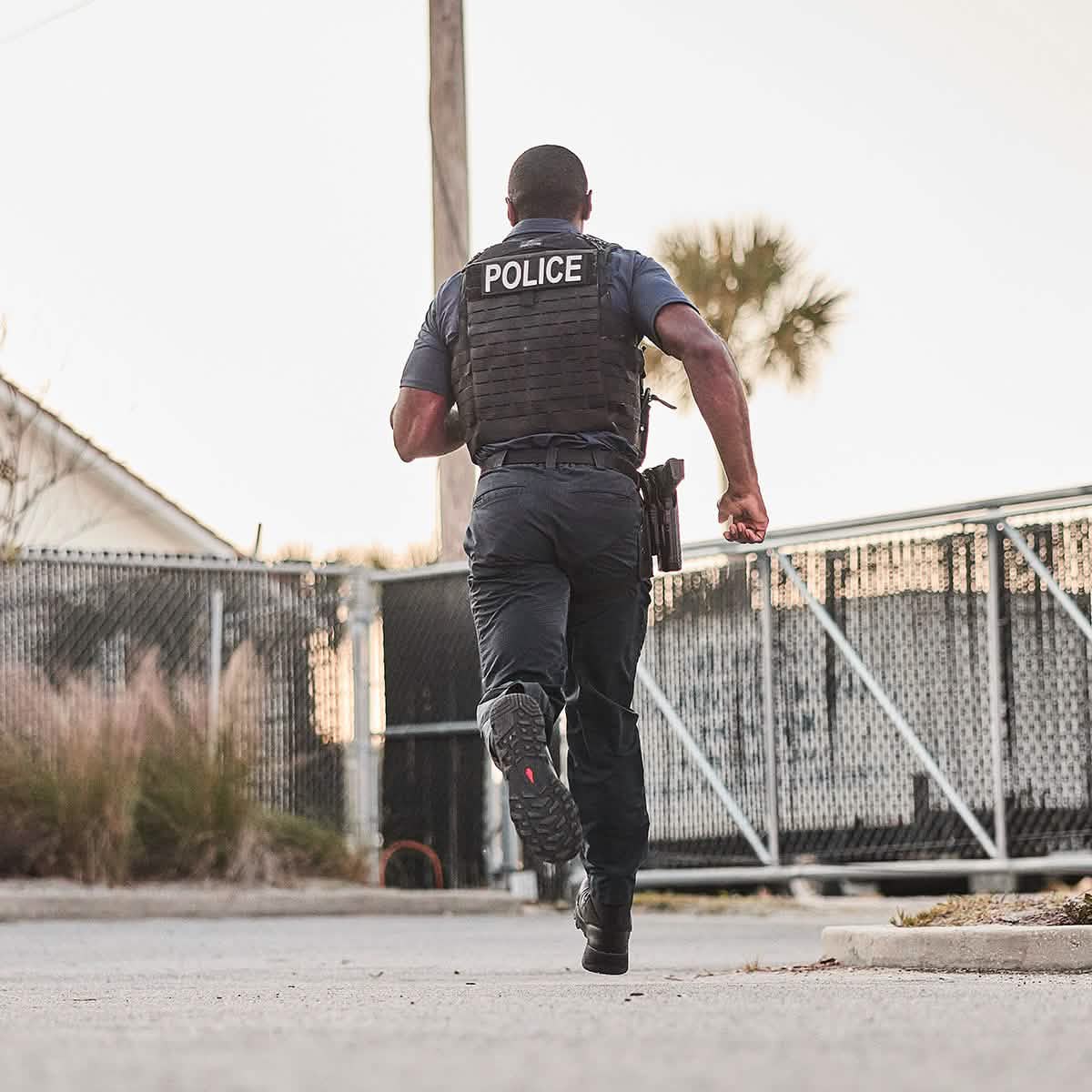 A police officer in a vest labeled POLICE dashes down a paved path wearing GORUCK's MACV-2 Safety Boot - High Top. The backdrop includes a chain-link fence, dry grass, and a palm tree against a clear sky, indicating an outdoor urban environment.