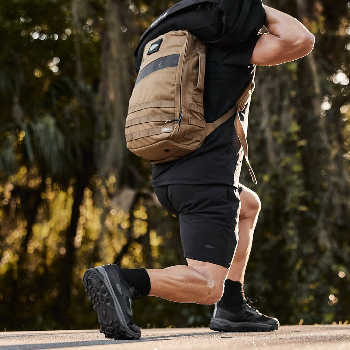 A person decked out in athletic gear is performing a lunge while carrying a brown backpack, echoing the essence of Special Forces training. Surrounded by trees and foliage, they're wearing comfortable black trail shoes and shorts from the Mackall - Black + Forged Iron collection by GORUCK.