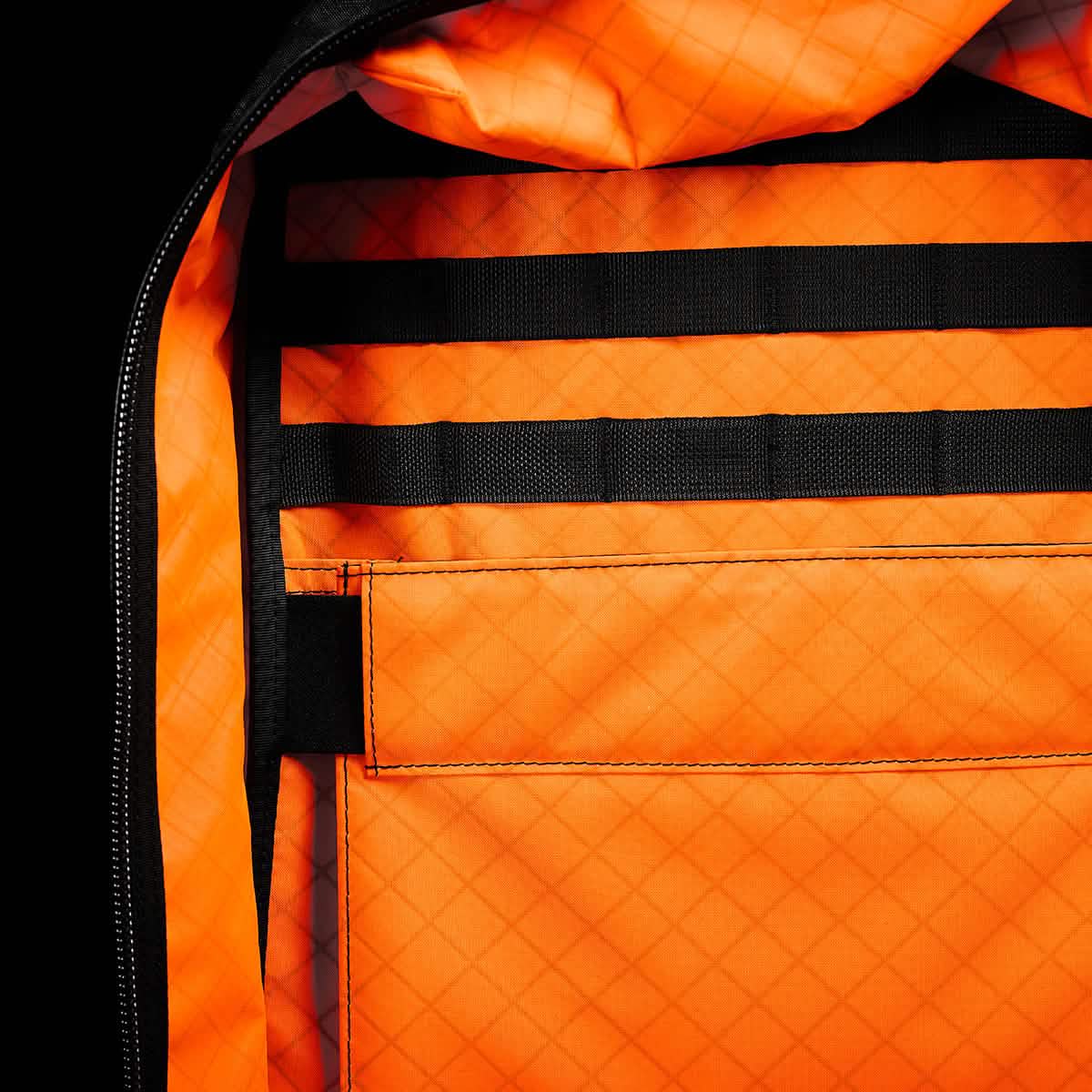 Close-up of an open GR2 USA x Carryology - Ultra Black backpack revealing a vivid orange, grid-patterned Challenge Sailcloth interior with black straps and a pocket.