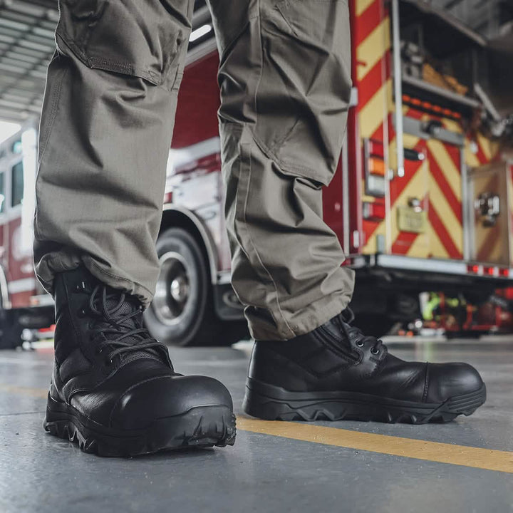 A person wearing khaki cargo pants and GORUCK's MACV-2 Safety Boot - High Top stands on a concrete floor. A red and yellow striped truck, possibly a fire truck, is visible in the background inside a garage.