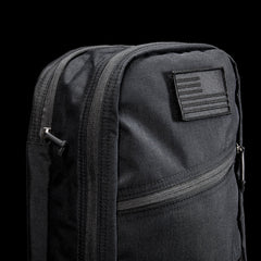 Close-up of the Bullet Ruck USA Throwback, a black daypack made with durable CORDURA, featuring a subdued American flag patch, multiple zippered compartments, and MOLLE webbing for versatile attachments.