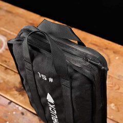 A black backpack with zippers and handles, adorned with the numeral 15 and a logo similar to RUCK, is tailored for adventurers seeking to carry their Sand Ruck Plates. Displayed on a rustic wooden surface, this GORUCK product comes with a lifetime guarantee for assurance.
