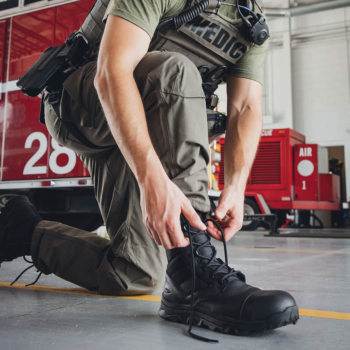 A medic in tactical gear kneels on the floor to tie the laces of his GORUCK MACV-2 Safety Boot - High Top. In the background, a red emergency vehicle is partially visible inside a garage, hinting at Special Forces standards.
