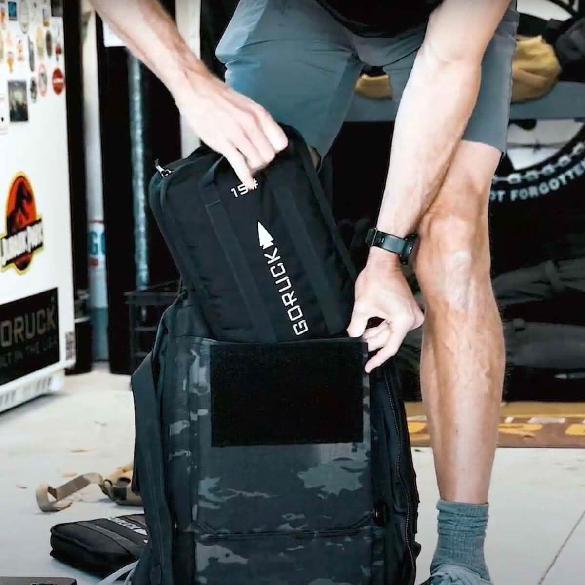 Person loading a Sand Ruck Plate Bundle into a black backpack, dressed in shorts and a watch—demonstrating must-have rucking gear in use.