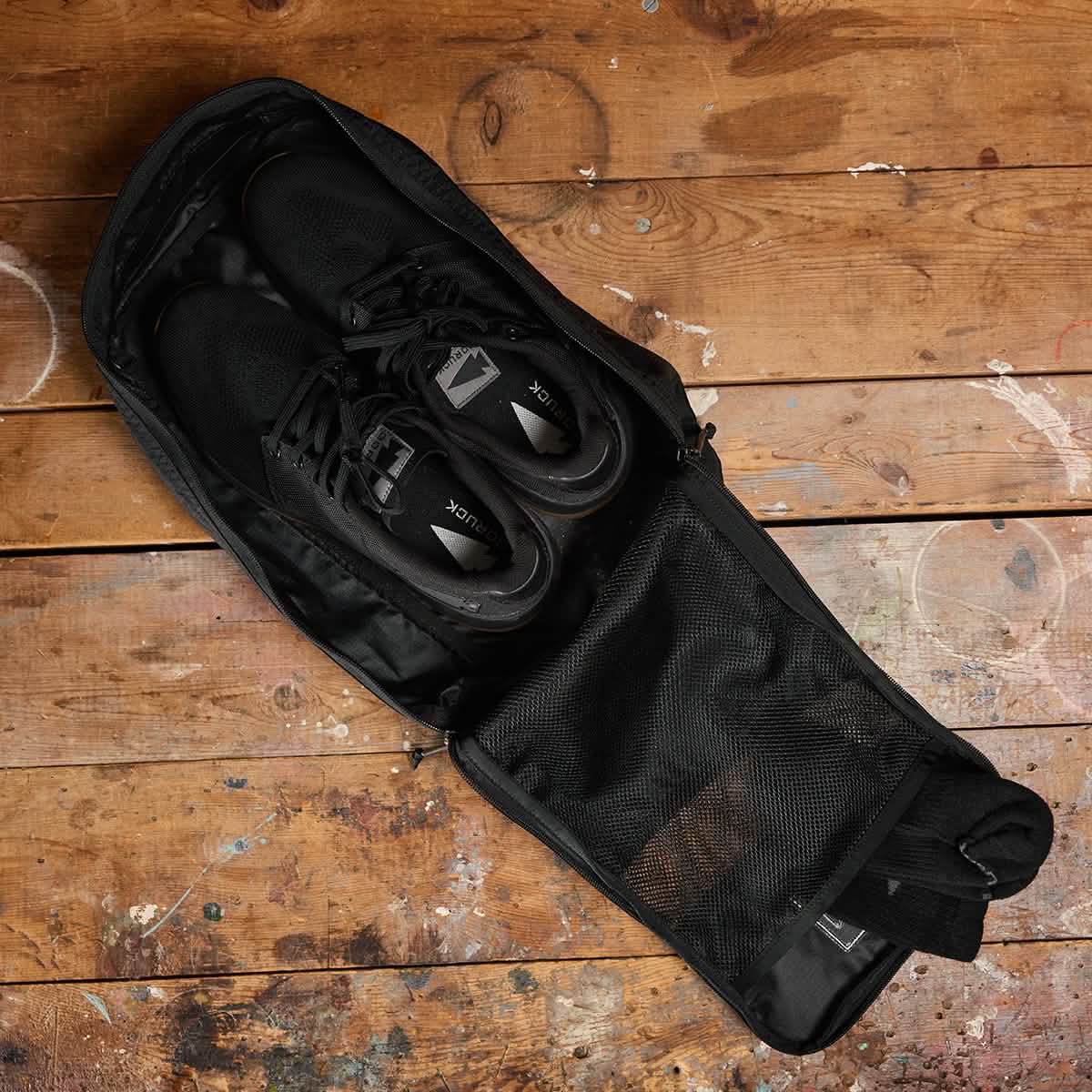 A pair of black sneakers is placed inside an open GORUCK Shoe Bag made from water-resistant Ripstop ROBIC® material, resting on a rustic wooden floor. The bag is partially zipped, revealing the neatly tucked sneakers inside. The floor features visible paint splatters and has a warm, worn texture.