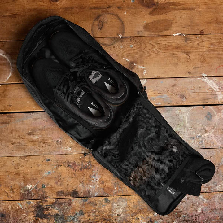 A pair of black sneakers is placed inside an open GORUCK Shoe Bag made from water-resistant Ripstop ROBIC® material, resting on a rustic wooden floor. The bag is partially zipped, revealing the neatly tucked sneakers inside. The floor features visible paint splatters and has a warm, worn texture.