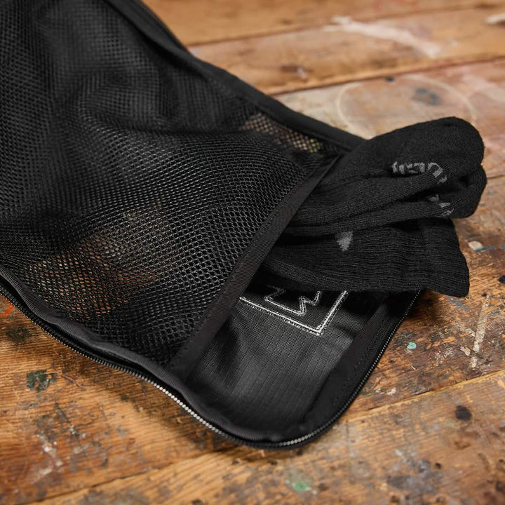 A Shoe Bag - Ripstop ROBIC® by GORUCK, in black mesh design and featuring a zipper closure, is partially open on a wooden surface, displaying several pairs of black socks inside.