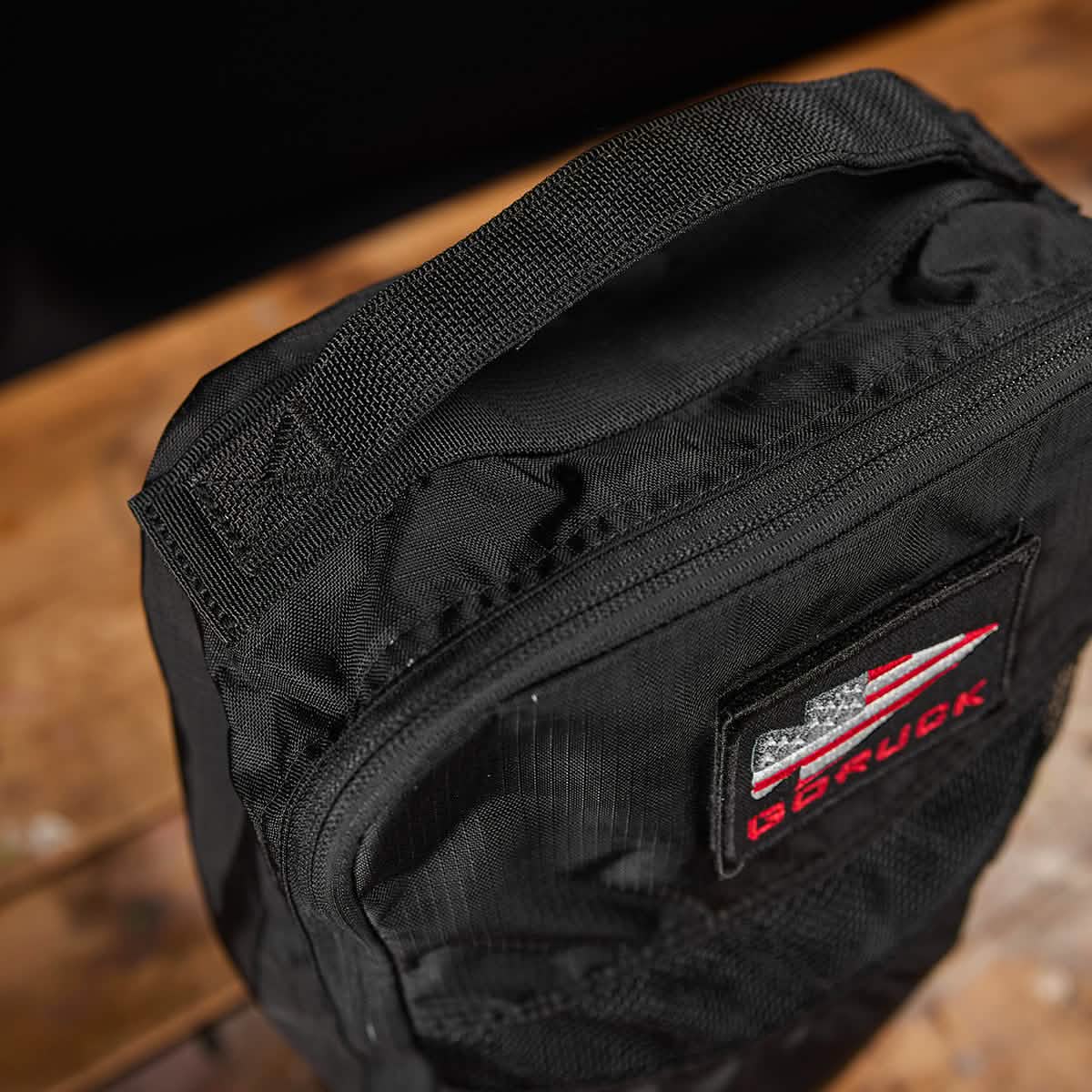 Close-up of a black shoe bag made from Ripstop ROBIC® material, featuring a patch with red and white stripes and stars. The brand name GORUCK is visible on the patch. Boasting a lifetime guarantee, this stylish bag offers peace of mind. It rests on a wooden surface, ready for your next adventure.
