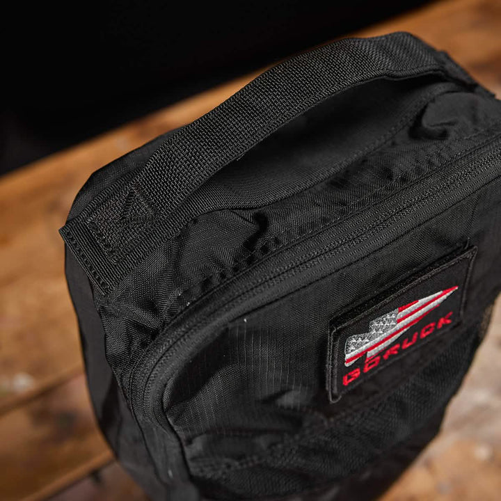 Close-up of a black shoe bag made from Ripstop ROBIC® material, featuring a patch with red and white stripes and stars. The brand name GORUCK is visible on the patch. Boasting a lifetime guarantee, this stylish bag offers peace of mind. It rests on a wooden surface, ready for your next adventure.