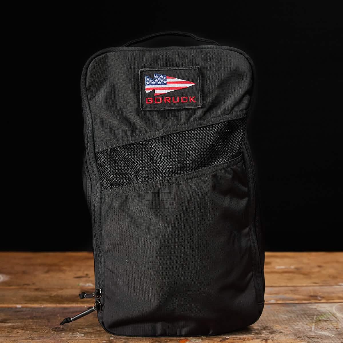 A black GORUCK backpack with a patch featuring an abstract American flag stands upright on a wooden surface against a black background. Made from water-resistant material, this design ensures durability and includes the Ripstop ROBIC® Shoe Bag, all backed by a lifetime guarantee.