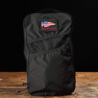 A black GORUCK backpack with a patch featuring an abstract American flag stands upright on a wooden surface against a black background. Made from water-resistant material, this design ensures durability and includes the Ripstop ROBIC® Shoe Bag, all backed by a lifetime guarantee.
