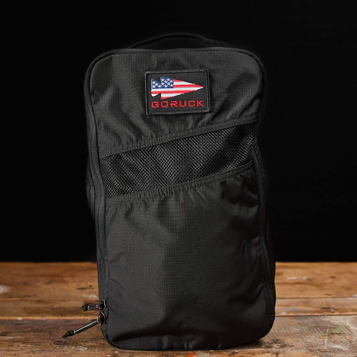 A black GORUCK backpack with a patch featuring an abstract American flag stands upright on a wooden surface against a black background. Made from water-resistant material, this design ensures durability and includes the Ripstop ROBIC® Shoe Bag, all backed by a lifetime guarantee.
