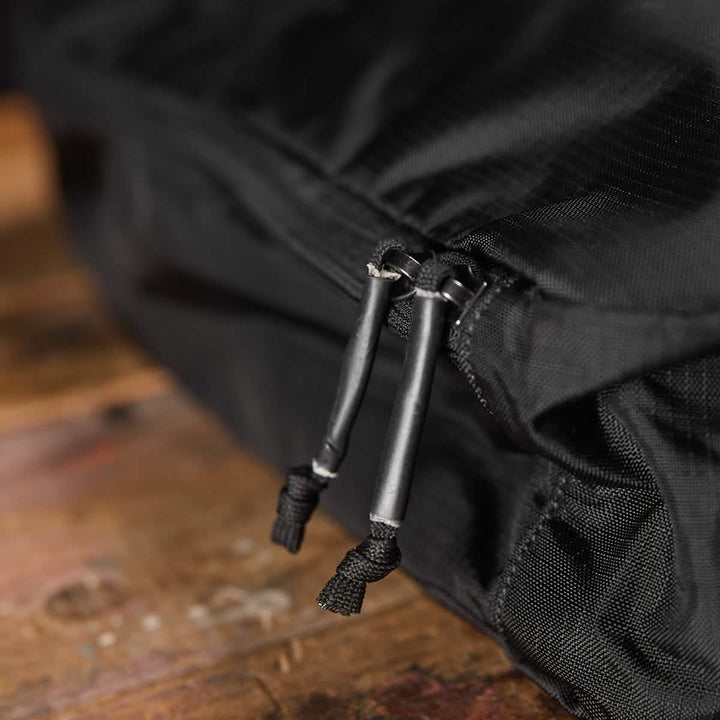 Close-up of the Shoe Bag - Ripstop ROBIC® by GORUCK, a black, water-resistant fabric bag with zippers featuring black pull cords for easy handling, placed on a wooden surface. The focus is on the zipper section while the slightly blurred background emphasizes this functional design.