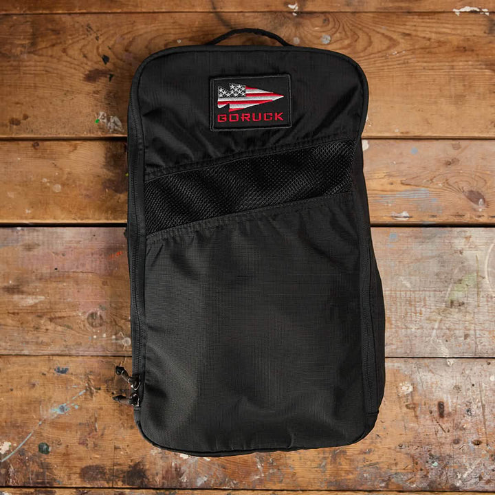 A Shoe Bag - Ripstop ROBIC® from GORUCK, with a patch showcasing the American flag and the brand name, sits on a wooden floor. This water-resistant bag features a mesh pocket on the front and comes with a lifetime guarantee, ensuring it is both reliable and rugged.