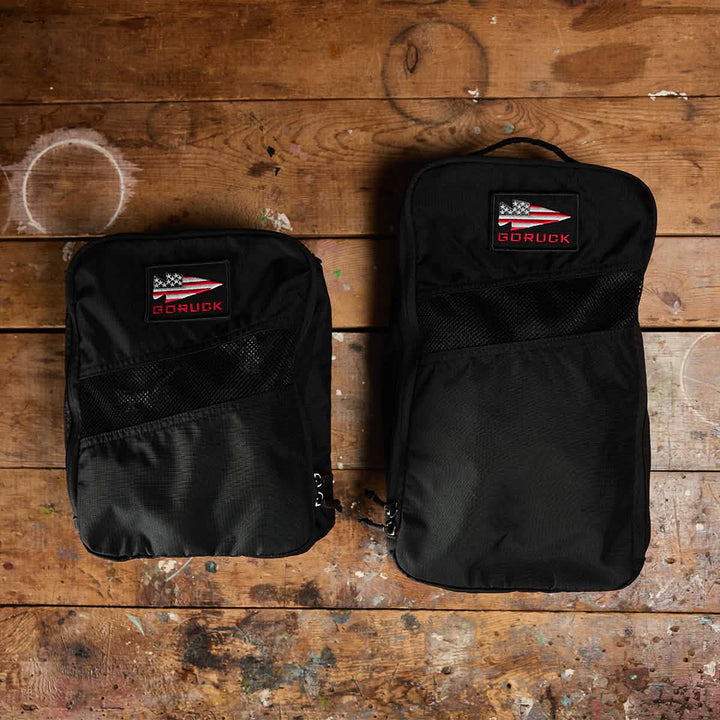 Two black GORUCK Shoe Bags made from Ripstop ROBIC® fabric, each featuring an American flag patch and differing in size, are placed side by side on a rustic wooden floor with paint stains. These water-resistant bags promise durability and are ready for any adventure or journey.