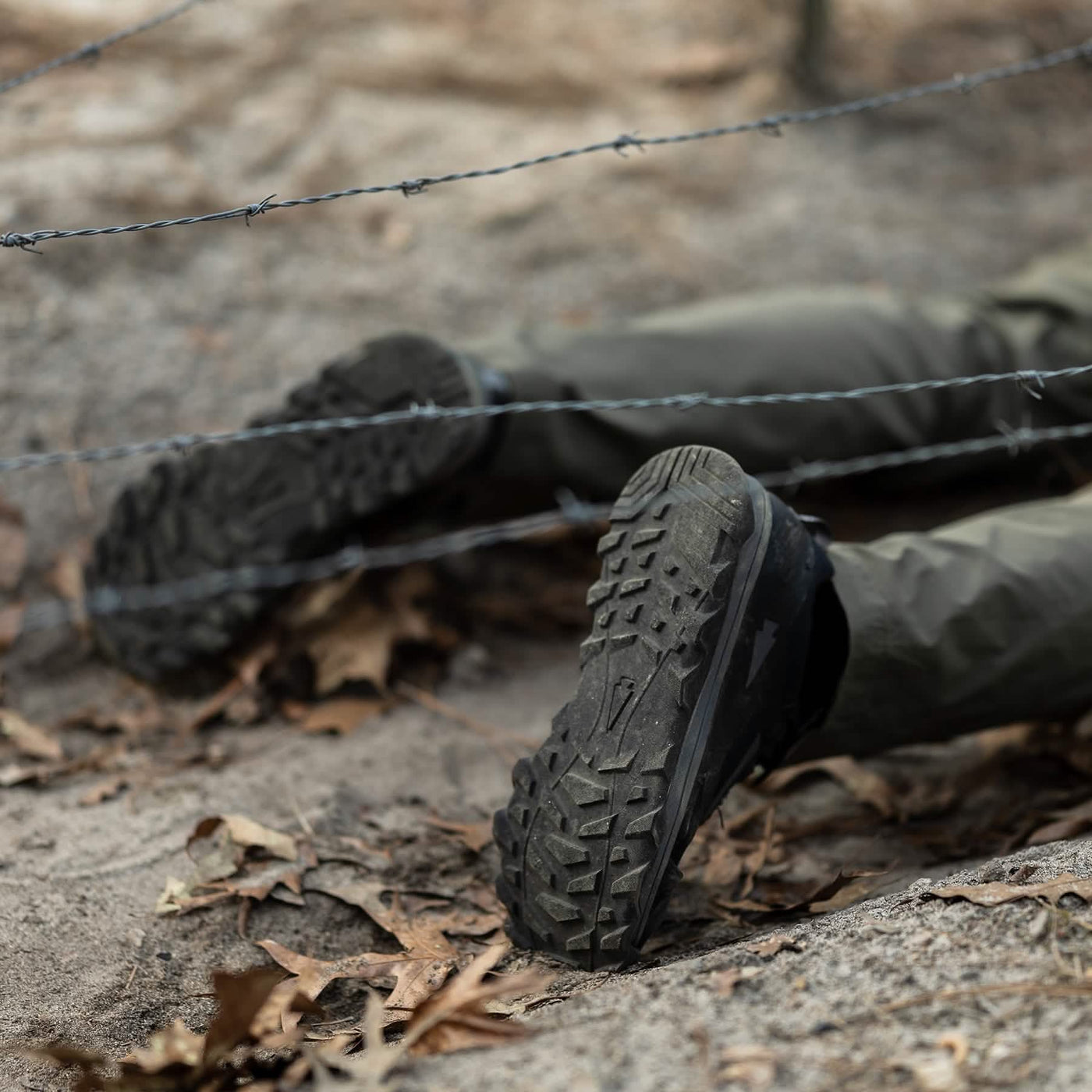 Close-up of a person lying on the ground beneath a barbed wire fence, their lower legs and feet visible in GORUCK Mackall shoes, in Black + Forged Iron color, paired with green pants. The terrain, evoking Special Forces training conditions, is strewn with dry leaves and dirt.