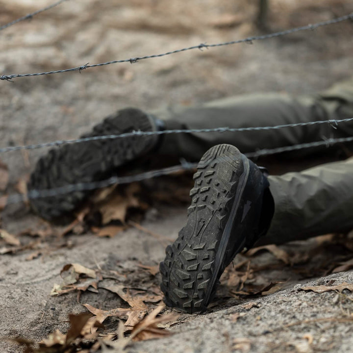 Close-up of a person lying on the ground beneath a barbed wire fence, their lower legs and feet visible in GORUCK Mackall shoes, in Black + Forged Iron color, paired with green pants. The terrain, evoking Special Forces training conditions, is strewn with dry leaves and dirt.