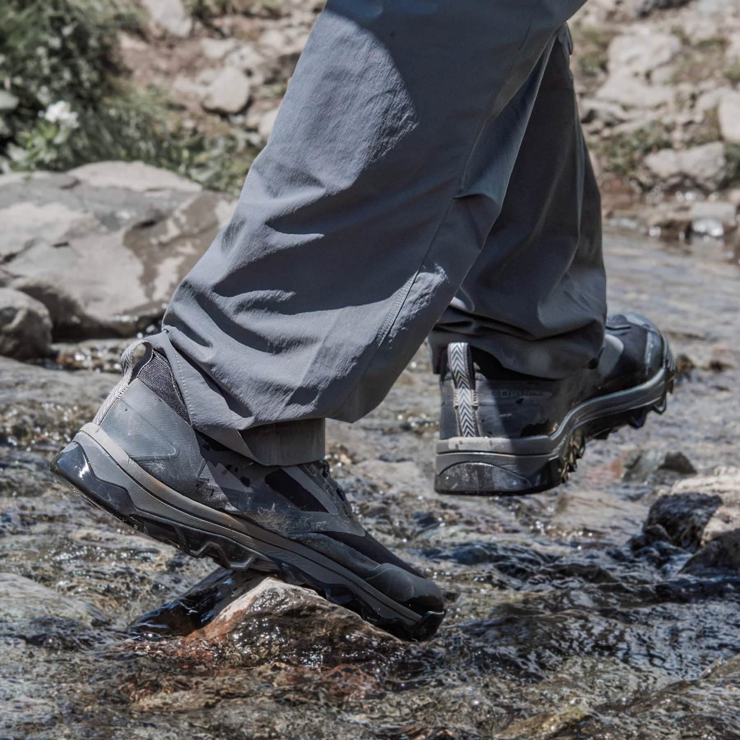 A person in gray pants crosses a small rocky stream, wearing GORUCK's Mackall shoes in Black + Forged Iron. The water is clear, revealing a few small rocks in this serene outdoor setting, reminiscent of Special Forces training grounds.